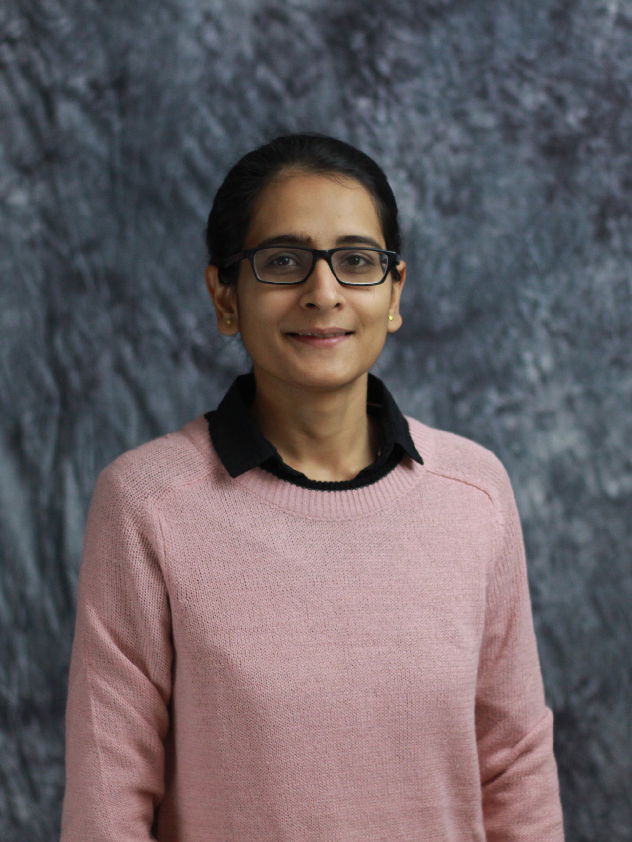 Headshot image of Mauli Pandey. She is wearing a pink sweater over a black shirt and standing in front of a grey background. She is smiling in the photo.